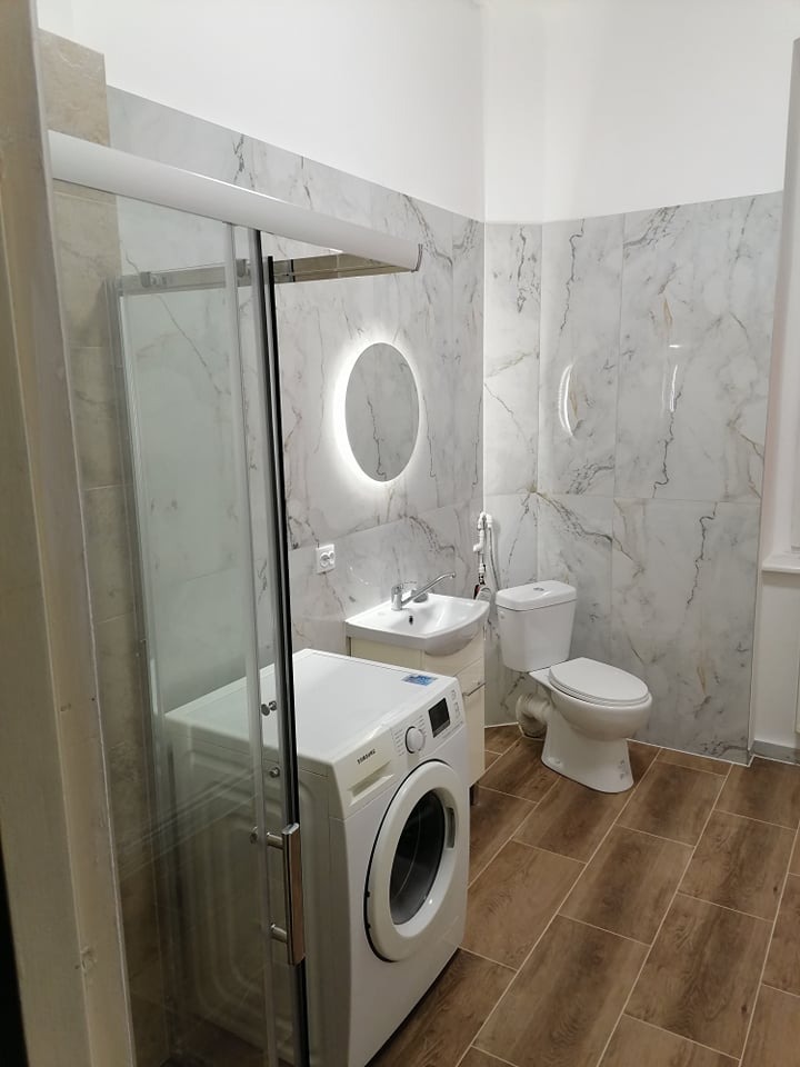 Wykończona łazienka z kabiną prysznicową, pralką, umywalką z szafką i toaletą, wyłożona płytkami imitującymi marmur i drewno, z okrągłym lustrem podświetlanym LED.