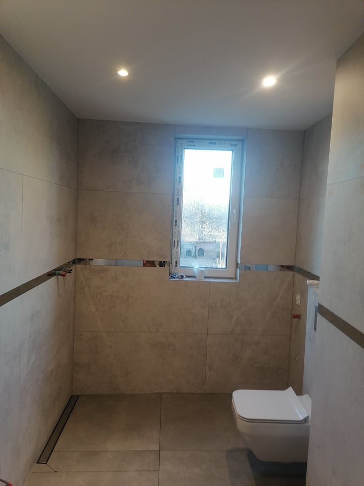 Wykończona łazienka z szarymi płytkami imitującymi kamień, minimalistyczna toaleta podwieszana, okno z widokiem na drzewa, chromowane listwy dekoracyjne i odpływ liniowy w podłodze.