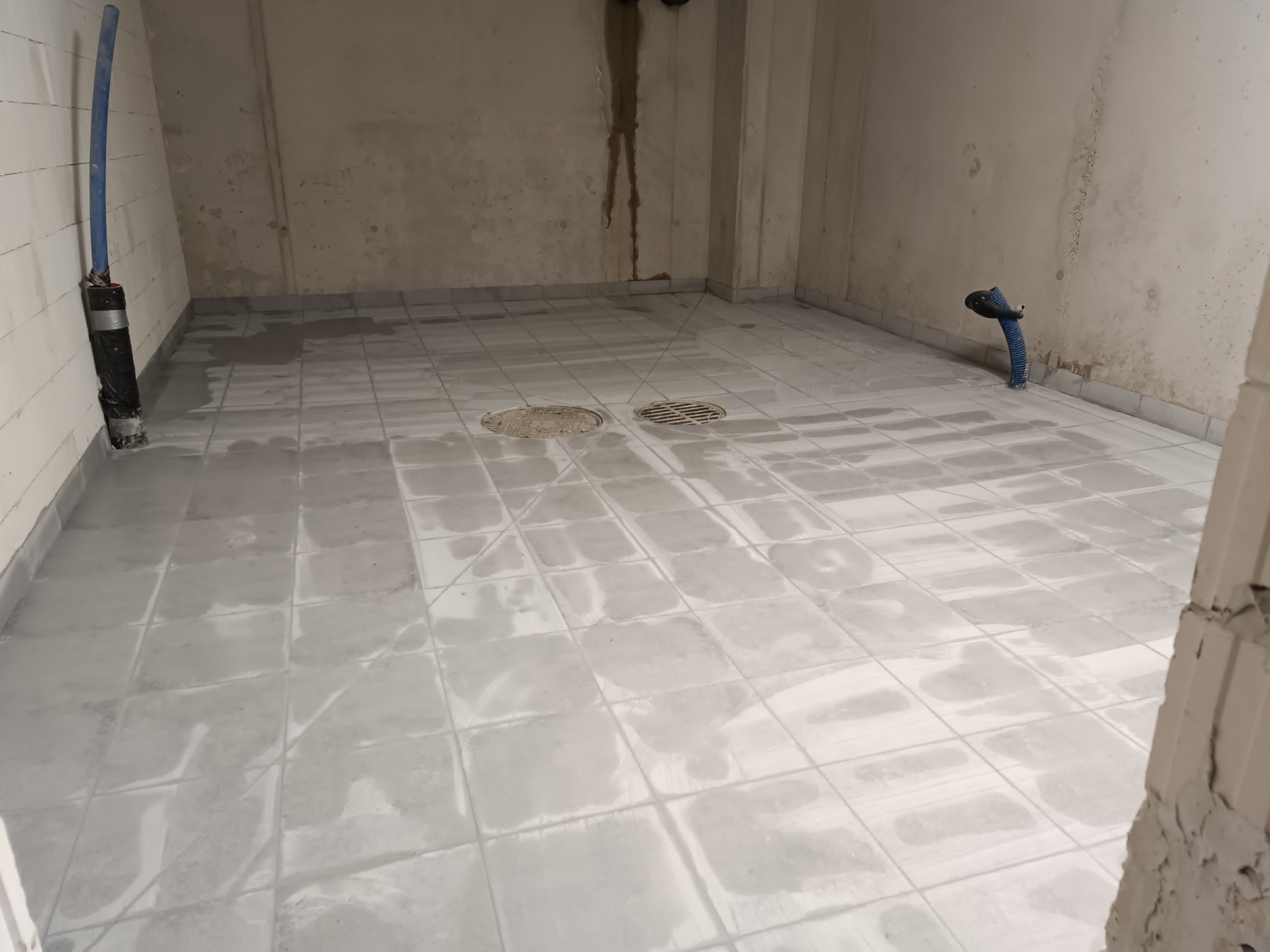 Świeżo wykafelkowana podłoga w łazience z odpływami, widoczne mokre fugi i fragmenty nieotynkowanych ścian.