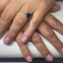 Dłonie kobiety z świeżo pomalowanymi paznokciami w odcieniu pudrowego różu, na serdecznym palcu srebrny pierścionek z szafirowym oczkiem, ułożone na białym tle.