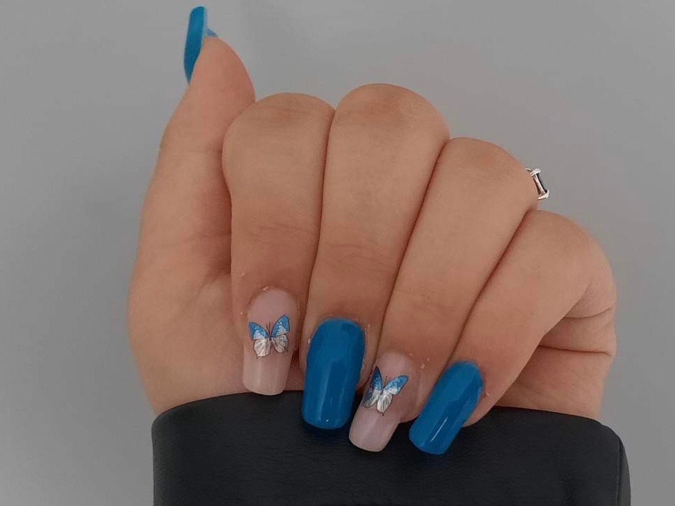 Dłoń z manicure hybrydowym w odcieniach błękitu, z motywem naklejonych, srebrno-błękitnych motyli na dwóch paznokciach, ujęcie z bliska.