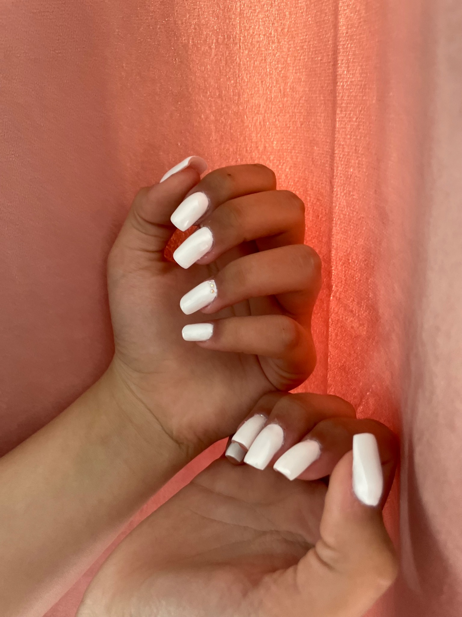 Dłonie z białym, kwadratowym manicure; jeden paznokieć zdobiony drobnymi cyrkoniami na tle różowej tkaniny.