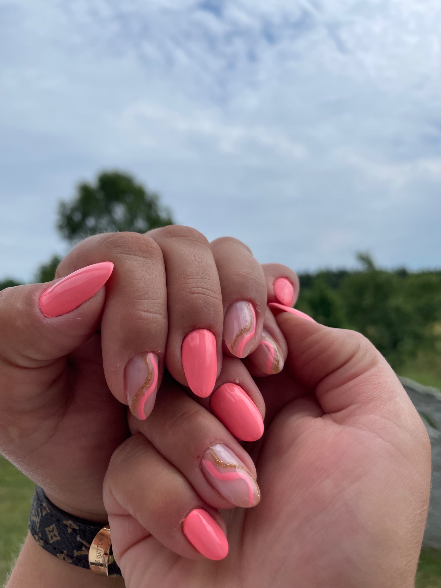 Dłonie z koralowymi paznokciami o migdałowym kształcie, jeden paznokieć zdobiony złotą linią, na tle błękitnego nieba i zieleni.