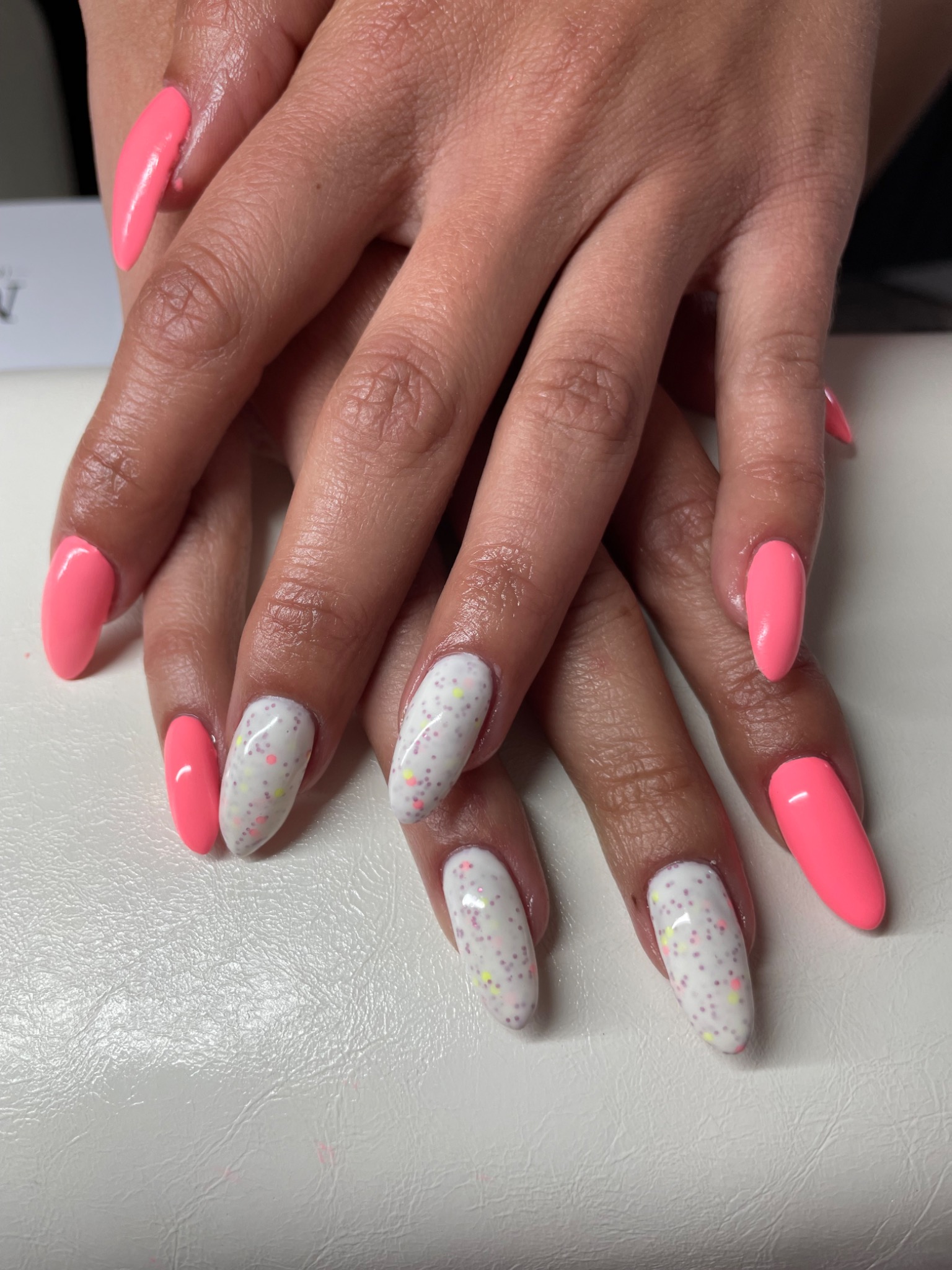 Dłonie z pomalowanymi paznokciami: niektóre w kolorze jasnego różu, inne białe z kolorowymi kropkami, na jasnym tle.