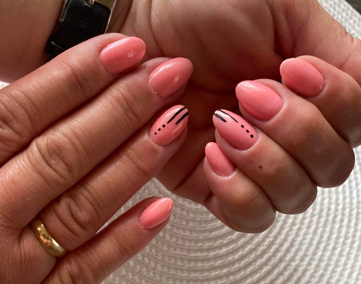 Ręce z paznokciami w odcieniu koralowym, zdobionymi czarnymi paskami i kropkami, pierścionek na palcu.