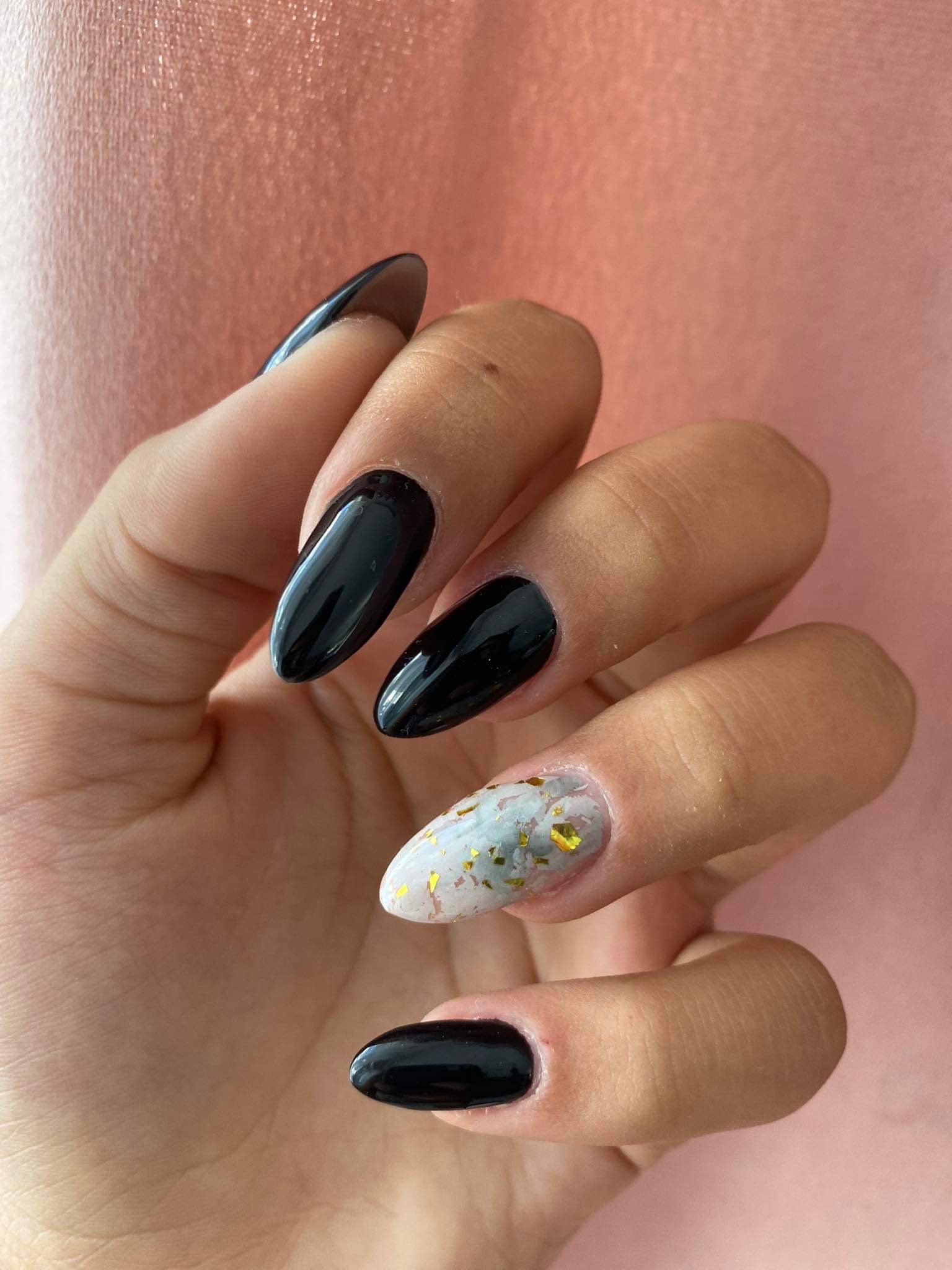 Dłoń z czarnymi, błyszczącymi paznokciami w kształcie migdała oraz jednym paznokciem z białym tłem i złotymi płatkami na tle brzoskwiniowym.