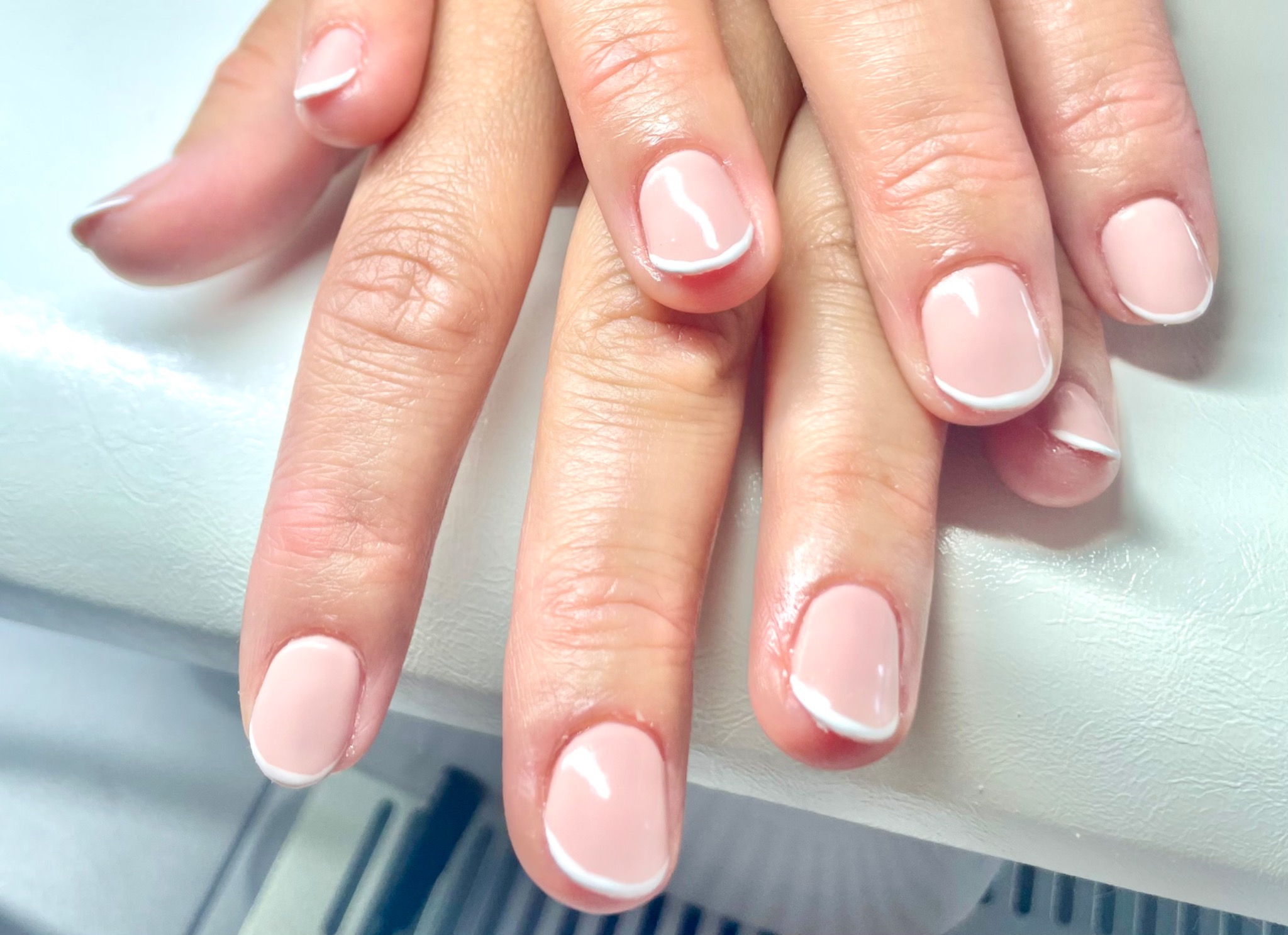 Delikatny, jasnoróżowy lakier z białym, cienkim paskiem na końcach paznokci u dłoni kobiety na białym tle.