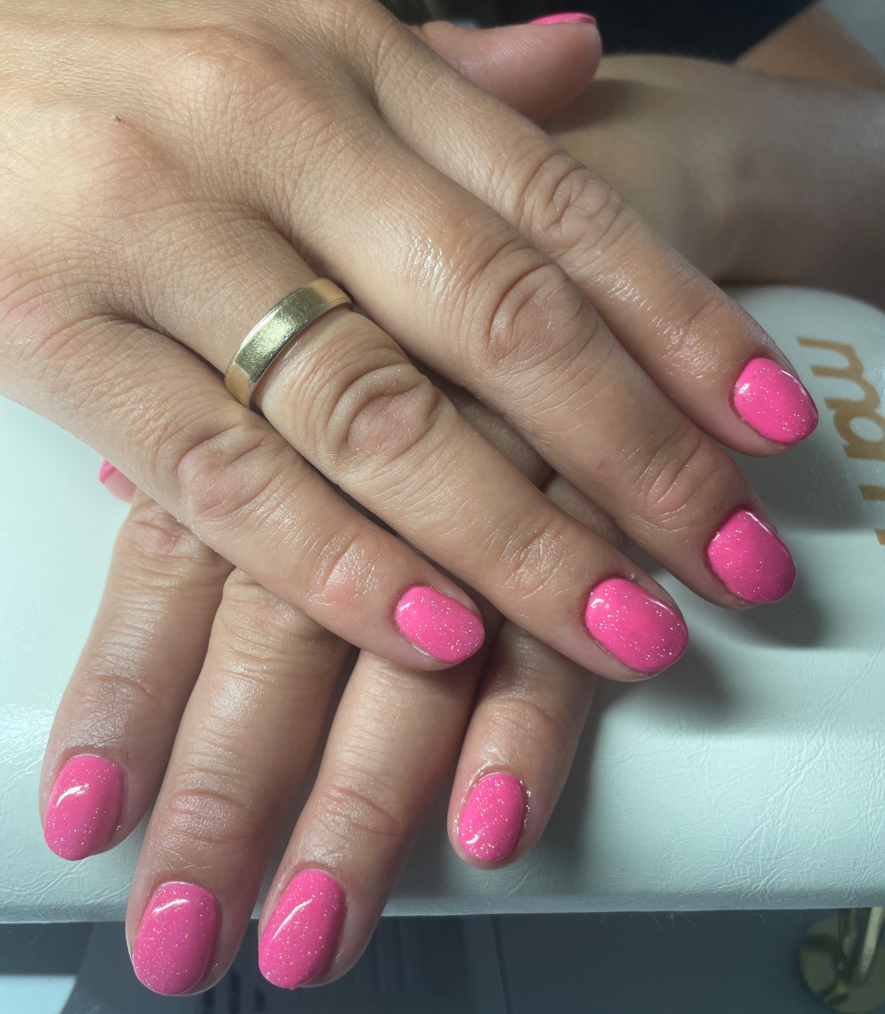 Dłonie kobiety z świeżo pomalowanymi paznokciami w odcieniu jaskrawego różu z delikatnym brokatem, na jednym palcu złoty pierścionek.