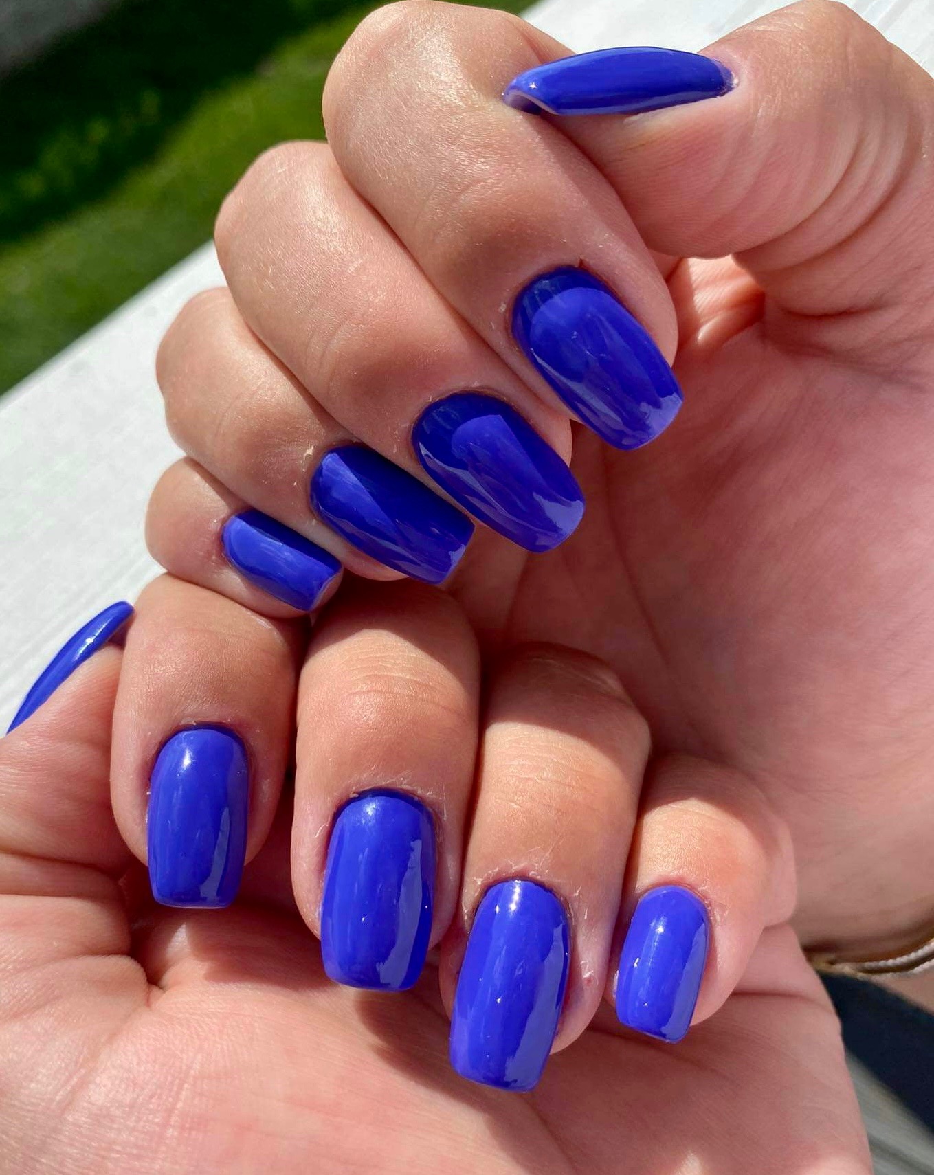 Dłonie z intensywnie niebieskim, błyszczącym lakierem na krótkich, zaokrąglonych paznokciach, sfotografowane w jasnym, naturalnym świetle.