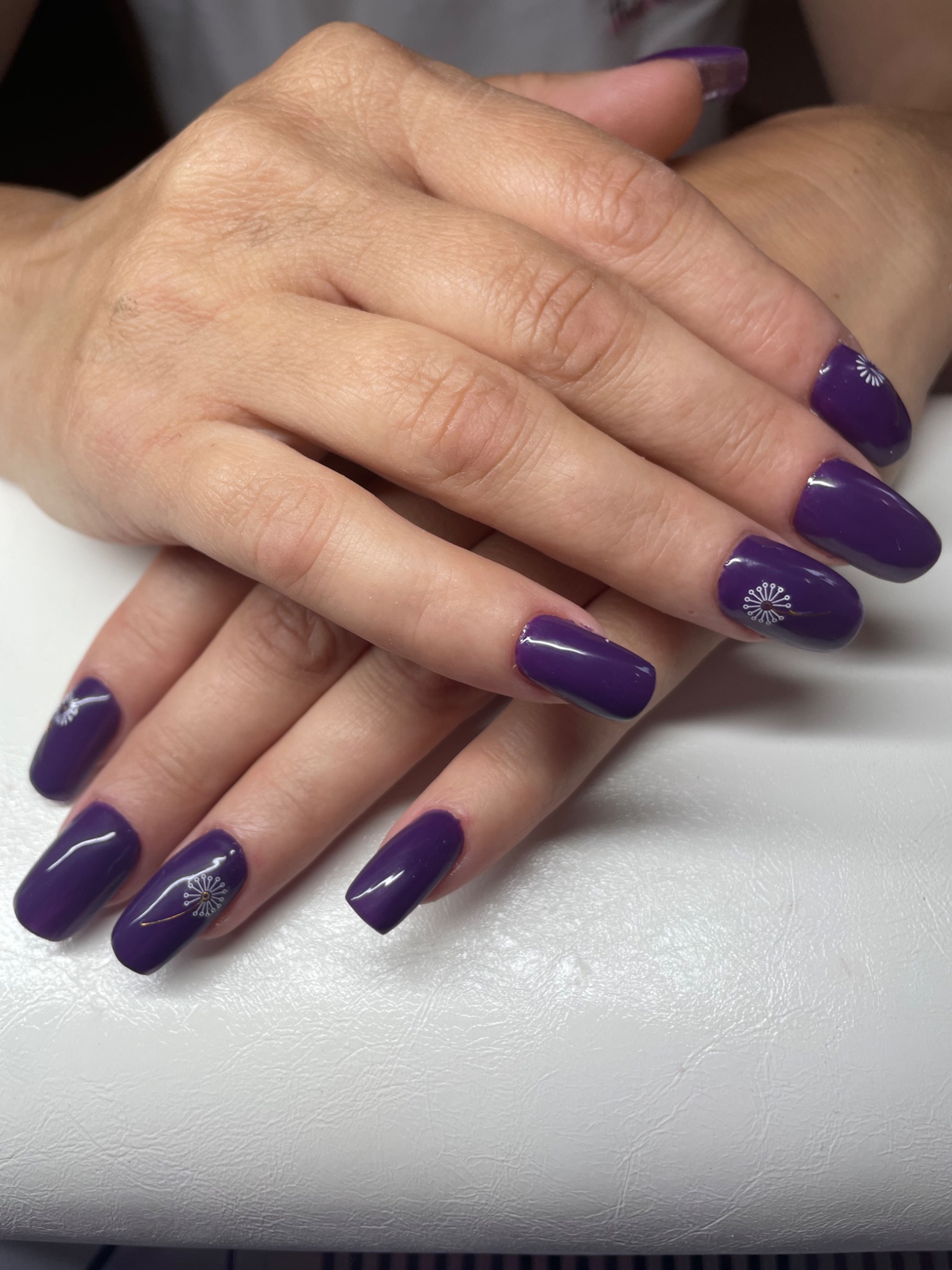 Dłonie z ciemnofioletowymi, błyszczącymi paznokciami w kształcie migdała, ozdobionymi drobnymi, białymi wzorami dmuchawca na kilku paznokciach, ułożone na białym tle.