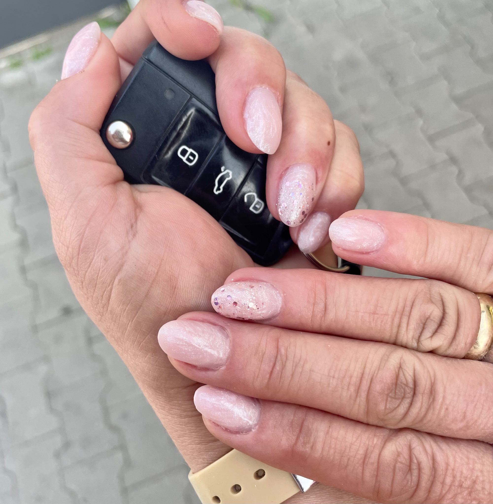 Jasnoróżowy manicure hybrydowy z brokatem na owalnych paznokciach, dłoń trzyma kluczyk samochodowy, w tle szara kostka brukowa.