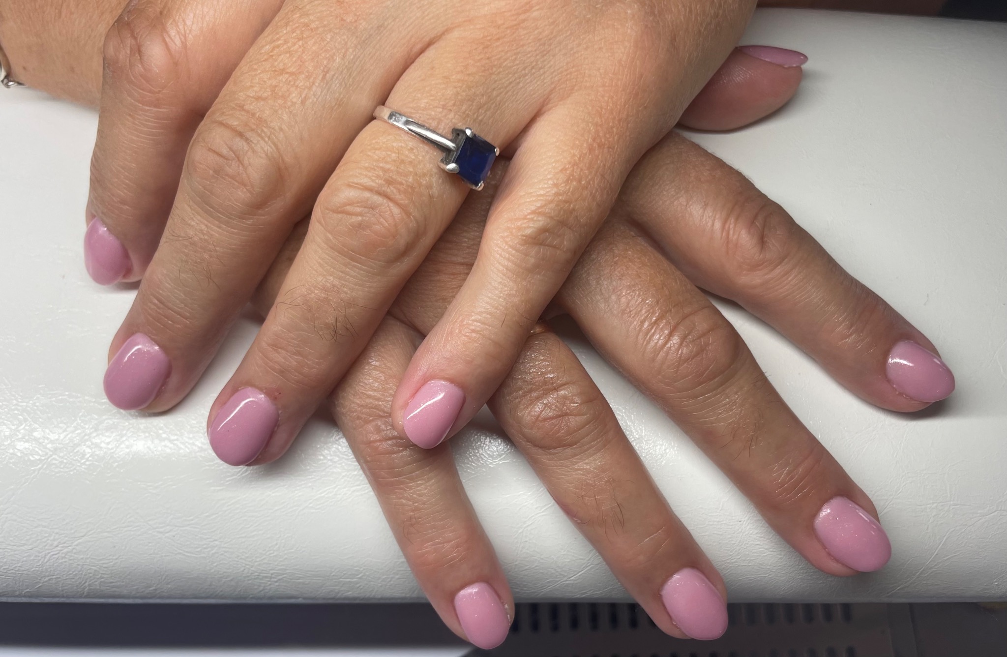 Dłonie kobiety z świeżo pomalowanymi paznokciami w odcieniu pudrowego różu, na serdecznym palcu srebrny pierścionek z szafirowym oczkiem, ułożone na białym tle.