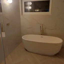 Nowoczesna łazienka z wolnostojącą białą wanną, złotą armaturą i marmurowymi płytkami w beżowych odcieniach z wyraźnym żyłowaniem, oświetlona lampą kulkową. Parkiet ułożony w jodełkę.