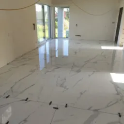 Białe, marmurowe płytki podłogowe z krzyżykami dystansowymi, świeżo ułożone w jasnym, pustym pokoju z dużymi oknami.