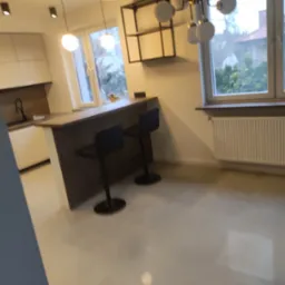 Nowoczesna kuchnia z barkiem i hokerami w minimalistycznym stylu, widok na okno z grzejnikiem, oświetlenie punktowe i wiszące lampy kuliste.