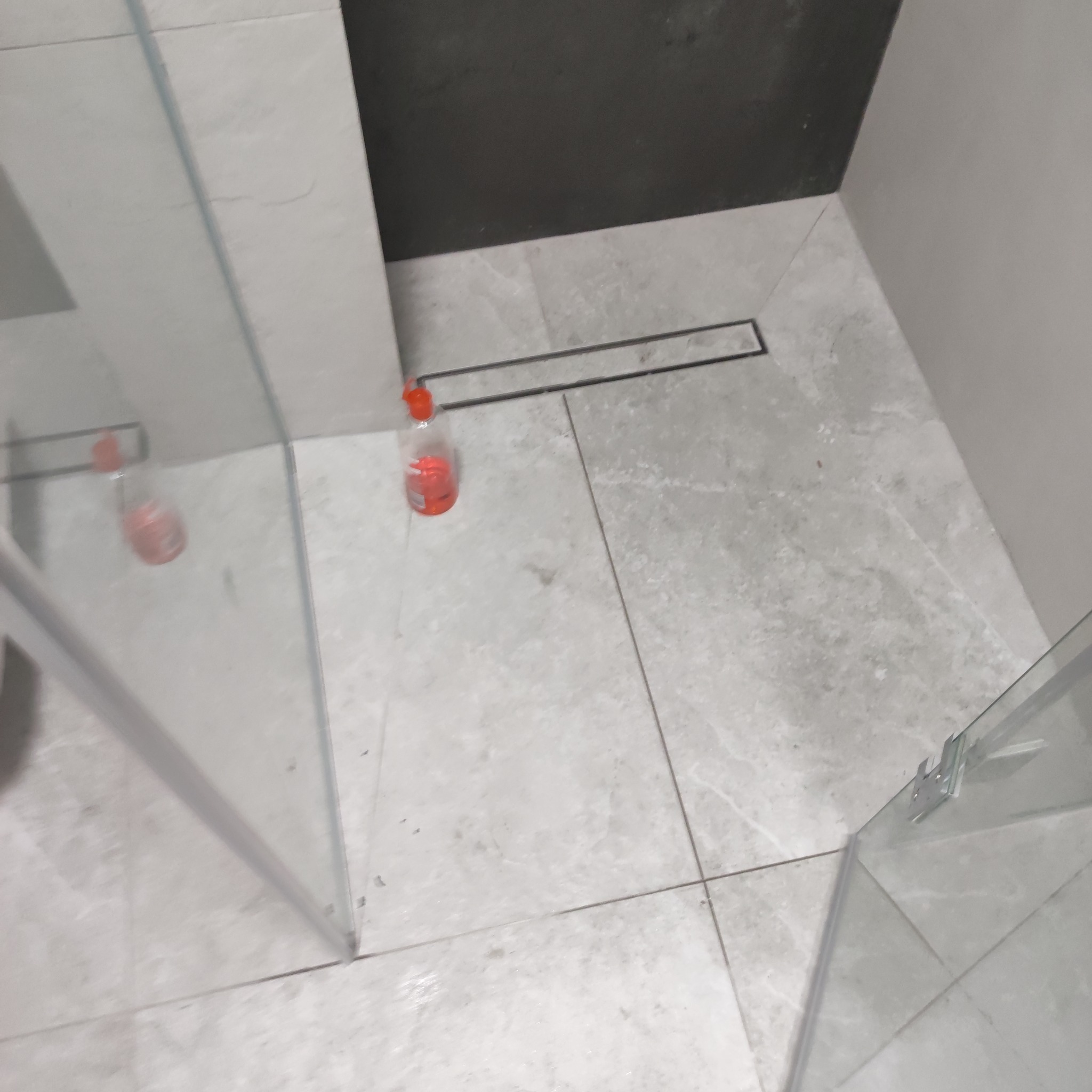 Ułożone jasnoszare płytki podłogowe w łazience z odpływem liniowym, widoczna szklana kabina prysznicowa i butelka z czerwonym płynem.