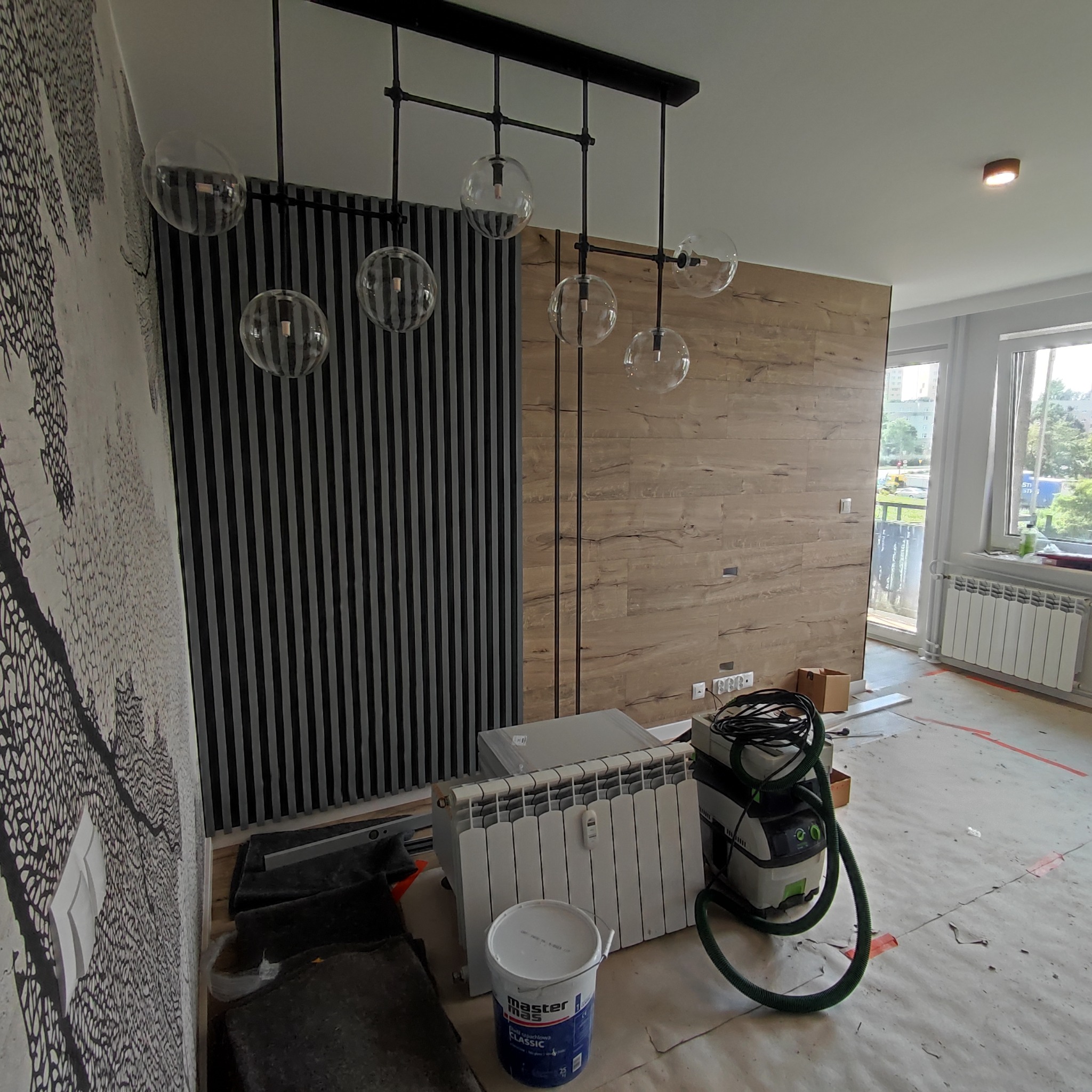 Wnętrze w trakcie prac wykończeniowych: widoczna ściana z tapetą w czarno-białe wzory, panelowa ściana z imitacją drewna, nowoczesna lampa z kulistymi kloszami, grzejnik, wiadro farby i odkurzacz...