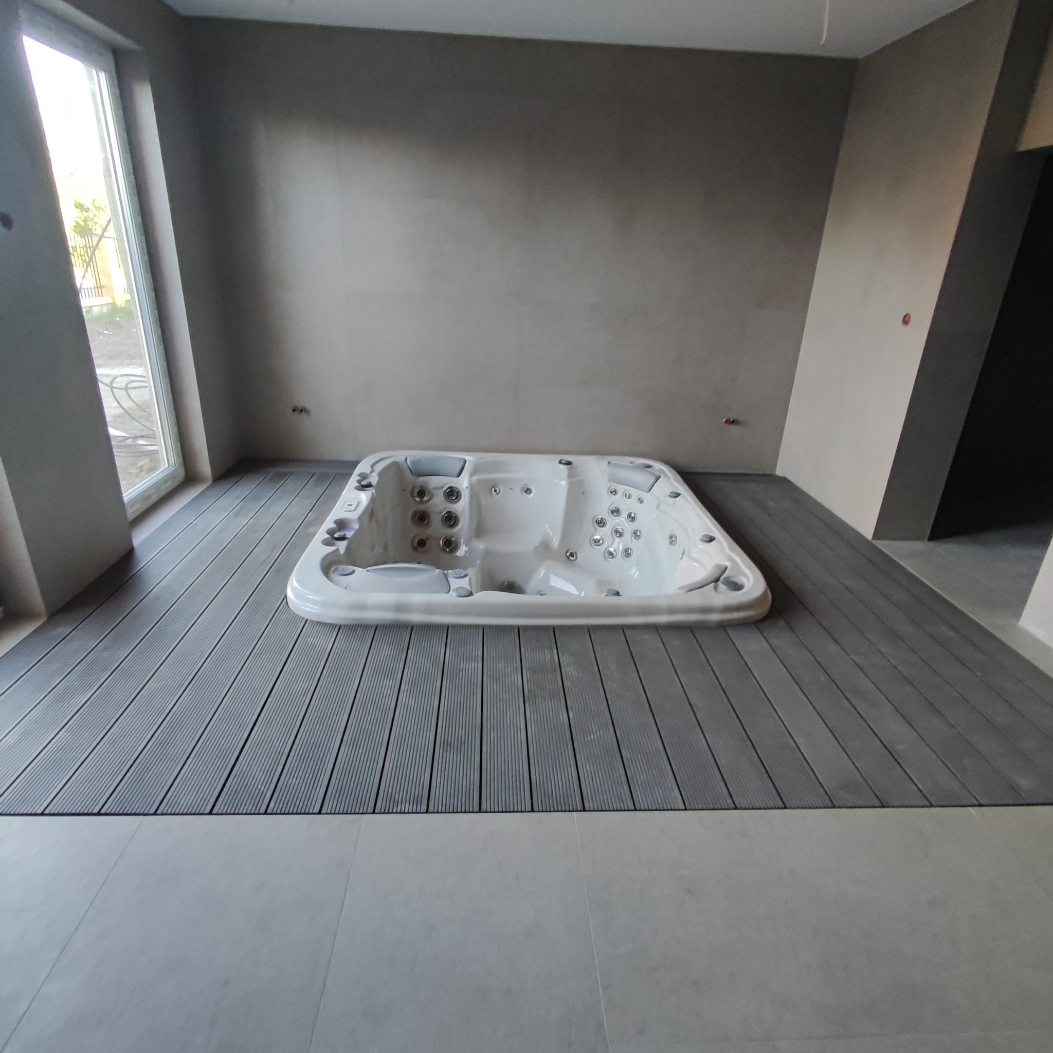 Biała wanna z hydromasażem na szarym, kompozytowym podeście w surowym wnętrzu z gładzią na ścianach i dużym oknem.