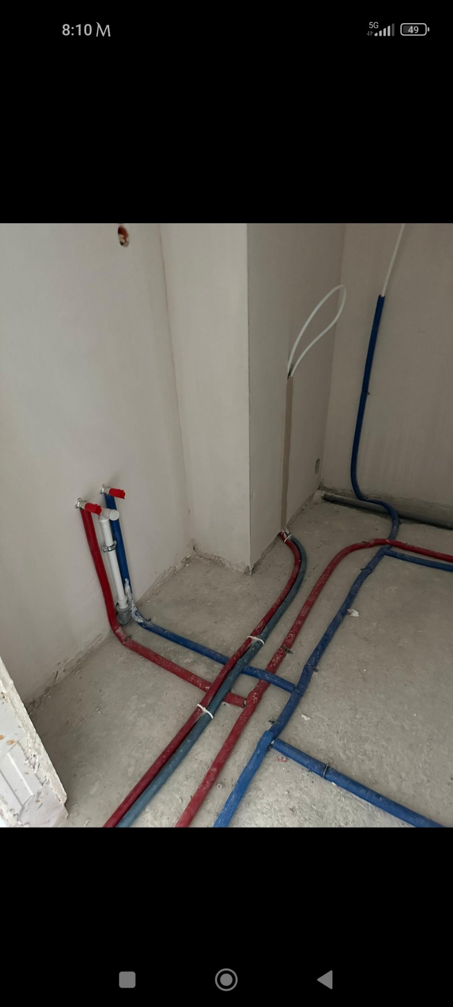 Świeżo rozprowadzone rury wodne, czerwone i niebieskie, na surowym betonie w narożniku łazienki, widoczne przyłącza do ściany.