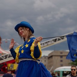 CINEMATOGRAPHY KONRAD GRADOWSKI - Uśmiechnięta kobieta w kostiumie z flagą UE na tle wesołego miasteczka. Nietypowy strój, niebieski kapelusz i brokat na twarzy. W tle karuzela i budynki.