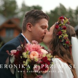 CINEMATOGRAPHY KONRAD GRADOWSKI - Ujęcie całującej się pary młodej w plenerze, z bukietem kwiatów i wiankiem na głowie panny młodej. W tle widoczny budynek z drewnianą architekturą.