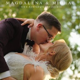 CINEMATOGRAPHY KONRAD GRADOWSKI - Pocałunek pary młodej w plenerze, ona w sukni z piórami, on w bordowym garniturze. Ujęcie z bliska, z widocznym tłem zieleni i nazwą fotografa u góry.