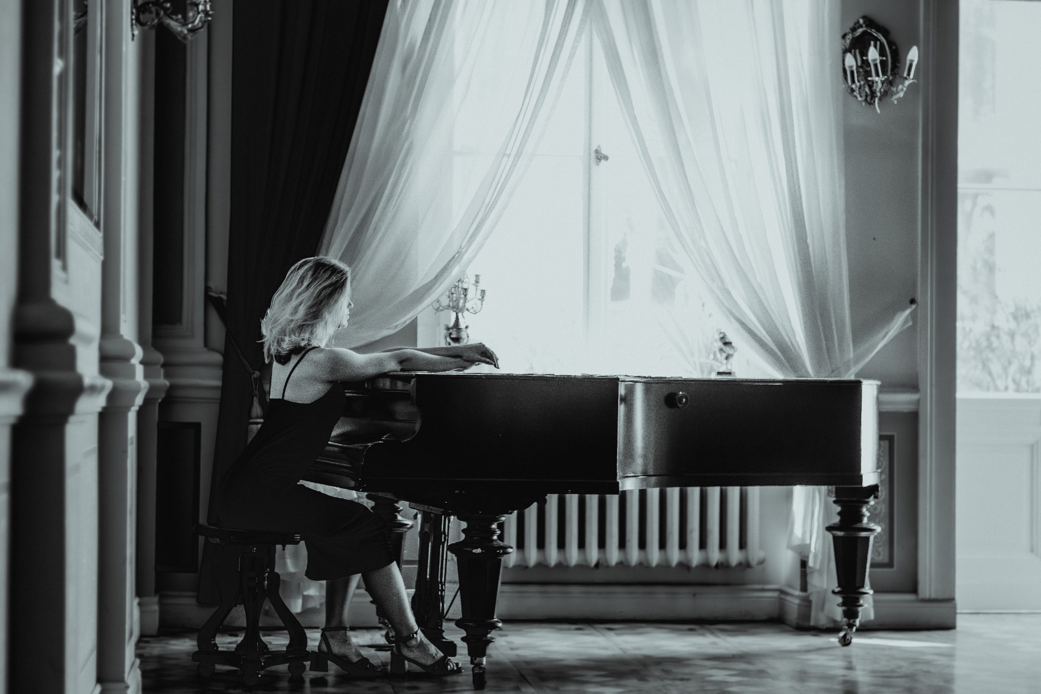 Kobieta w sukience siedzi przy fortepianie w jasnym pokoju z kolumnami i wysokim oknem, na fortepianie stoi świecznik.