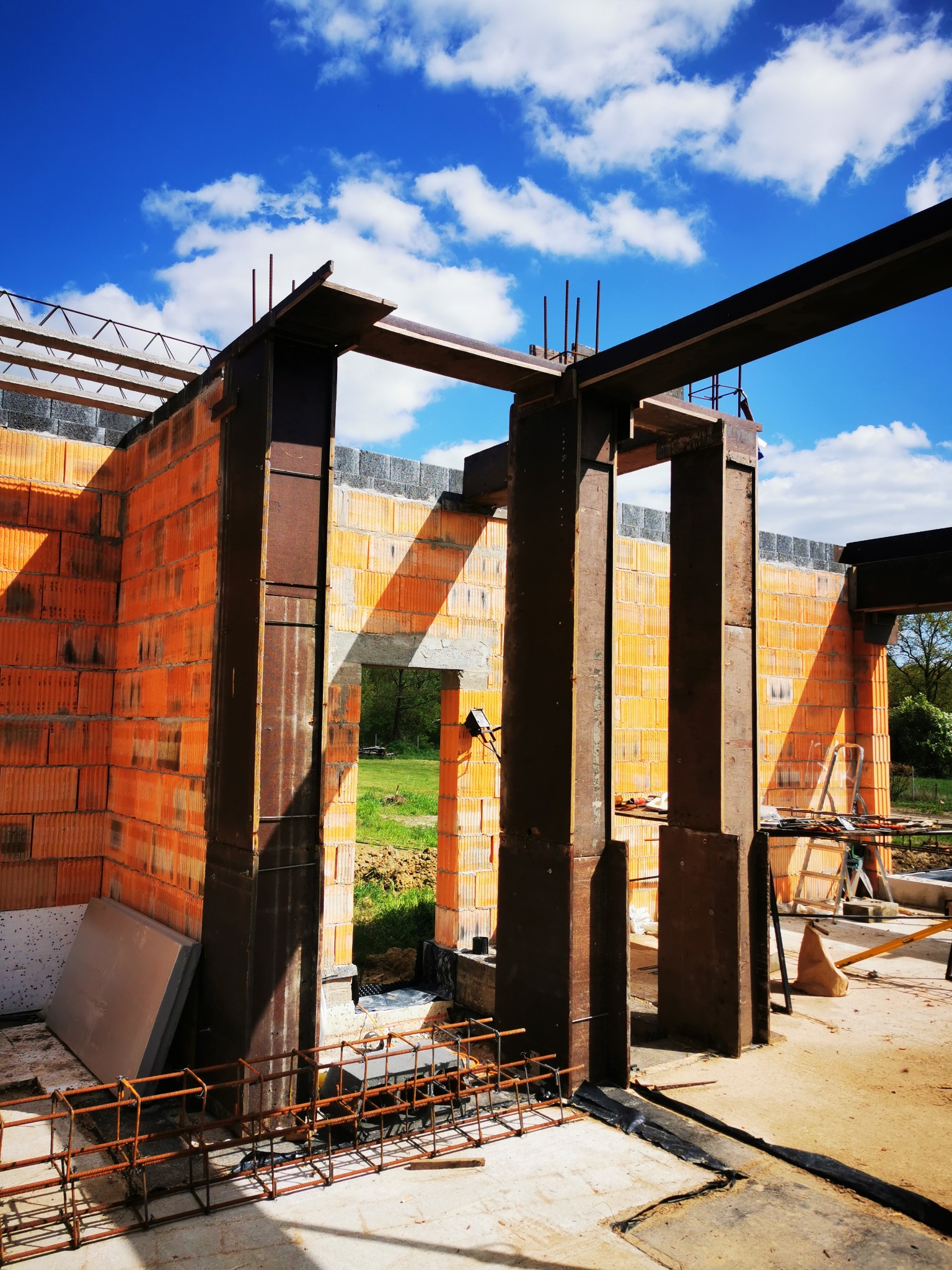 Wznoszenie muru z czerwonej cegły z elementami konstrukcji stalowej, widok na niebo z chmurami, w tle zieleń i częściowo wykończona podłoga z elementami zbrojenia.
