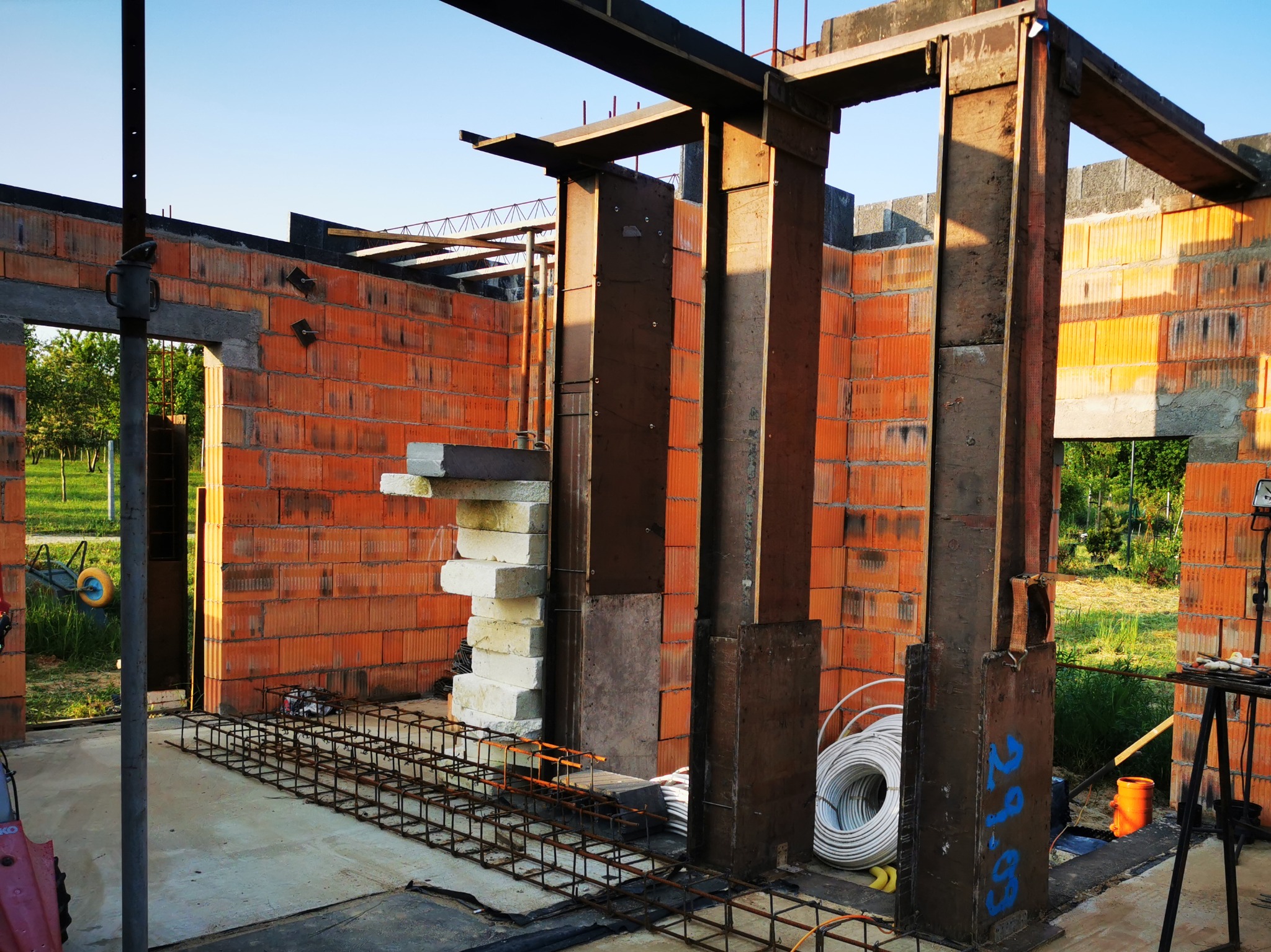 Wnętrze budynku w trakcie budowy, widoczne ściany z czerwonej cegły, stalowe słupy podtrzymujące konstrukcję, ułożone zbrojenie pod wylewkę oraz stos białych bloczków. Na zewnątrz widoczna zieleń.
