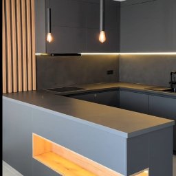 Piomax Piotr Kamiński - Nowoczesna kuchnia w odcieniach szarości z drewnianymi akcentami: listwy na suficie, podświetlona wnęka w wyspie kuchennej i oświetlenie LED pod szafkami.