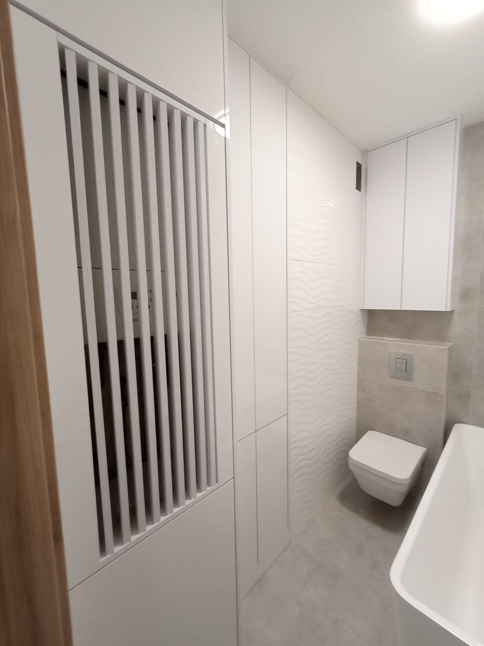 Biała szafa wnękowa z lamelami maskującymi licznik w łazience w Kościerzynie, dopasowana do minimalistycznego wystroju z sanitariatami w tle.