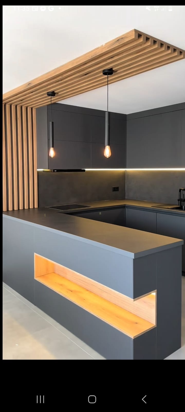 Nowoczesna kuchnia w odcieniach szarości z drewnianymi akcentami: listwy na suficie, podświetlona wnęka w wyspie kuchennej i oświetlenie LED pod szafkami.