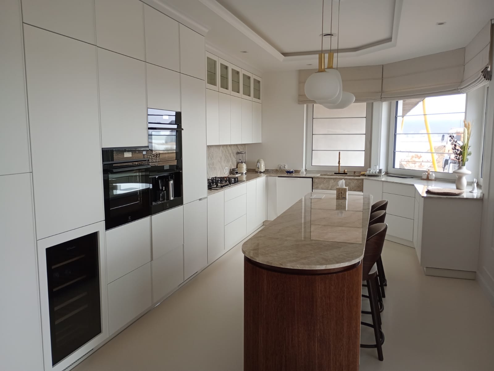 Nowoczesna kuchnia z białymi, lakierowanymi frontami, wyspą z marmurowym blatem i dekoracyjnym, żłobionym wykończeniem oraz wbudowanym sprzętem AGD, w tym piekarnikiem i chłodziarką do wina.