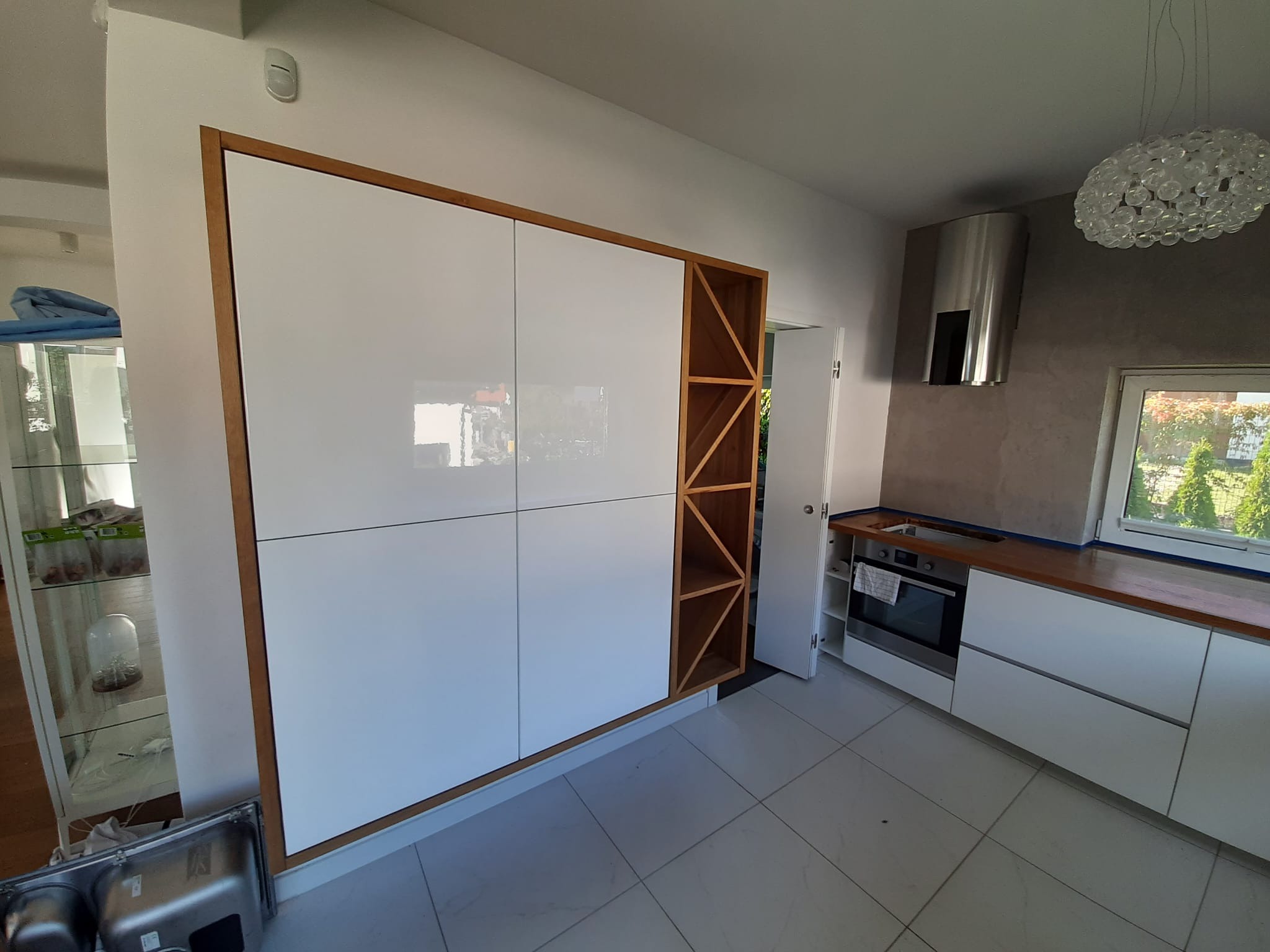 Biała kuchnia na wymiar z drewnianymi akcentami, obejmująca szafę wnękową z półkami na wino i zabudowę kuchenną z drewnianym blatem oraz srebrnym okapem.