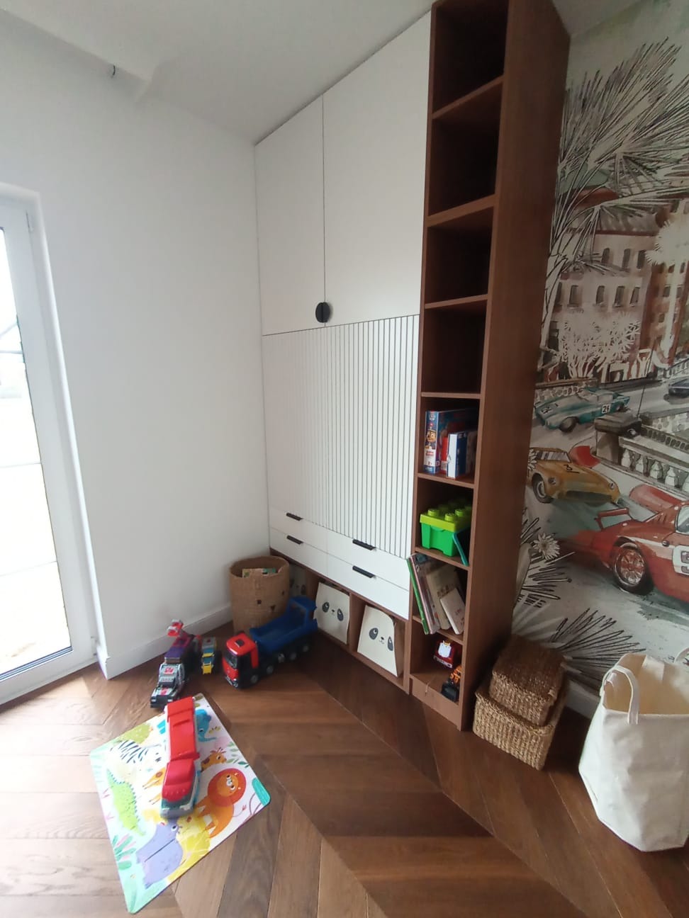 Biało-brązowa szafa w pokoju dziecięcym z zabawkami na podłodze i półkach, na ścianie fototapeta z motywem wyścigowym.