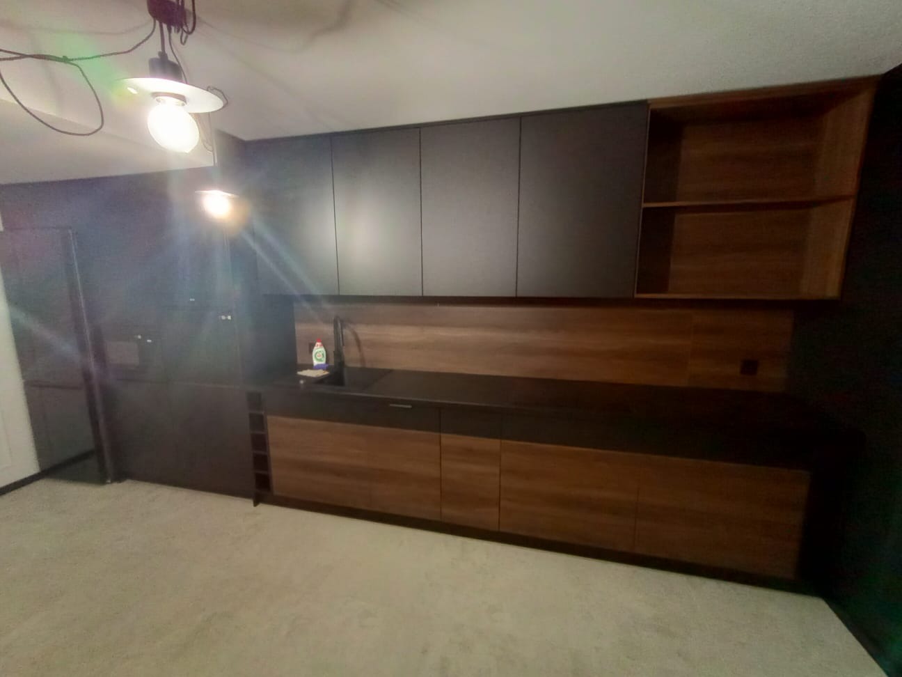 Nowoczesna zabudowa kuchenna z ciemnymi, gładkimi frontami szafek górnych i drewnopodobnymi frontami dolnymi, z otwartą półką po prawej stronie i widocznym oświetleniem sufitowym.