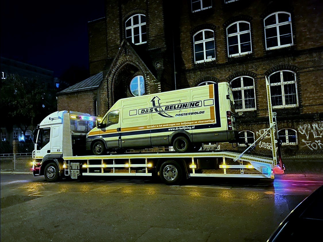 Laweta przewozi białą furgonetkę z logo D&S Belijning na tle ceglanego budynku w nocy.