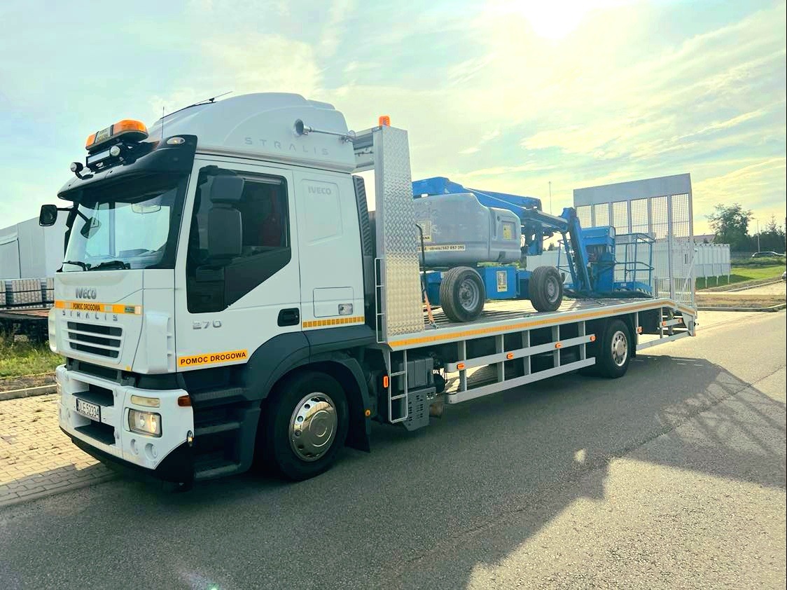 Biała ciężarówka Iveco Stralis z platformą transportową, przewożąca niebieski podnośnik koszowy. Widoczny napis 'Pomoc Drogowa' na boku kabiny.