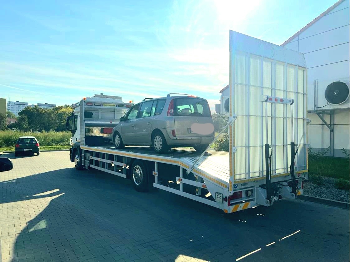 Szara osobówka Renault Espace na białej lawecie z pionową osłoną przeciwwiatrową, zaparkowanej na brukowanym placu w słoneczny dzień, z budynkiem w tle.