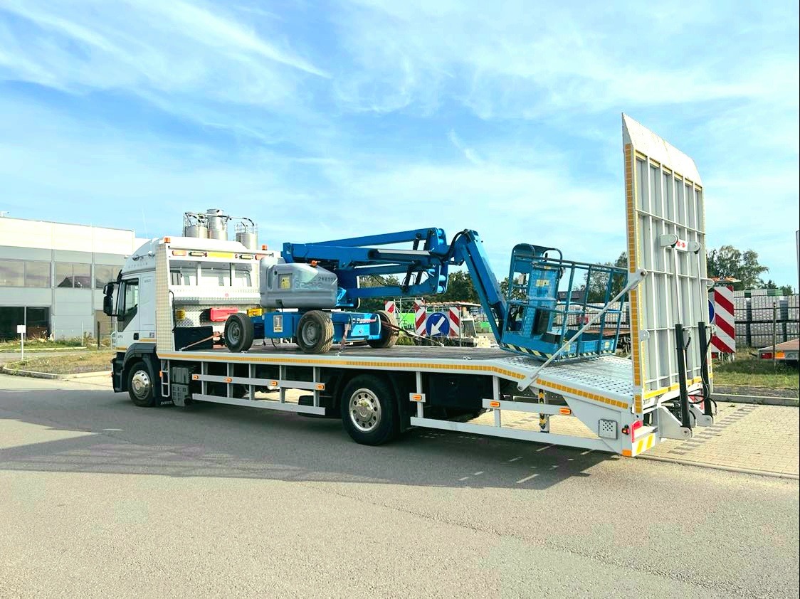 Laweta z niebieskim podnośnikiem koszowym na platformie, zabezpieczonym pionową osłoną z tyłu, zaparkowana na asfaltowej drodze w słoneczny dzień.