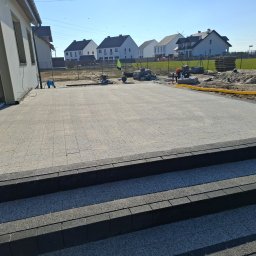 Piotr Wrzeszykowski - Ułożona jasnoszara kostka brukowa przed nowym domem, widoczne schody z ciemnej kostki, w tle prace wykończeniowe i hałda piasku, słoneczny dzień.