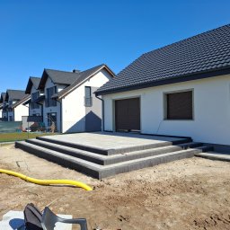 Piotr Wrzeszykowski - Nowoczesny taras z szarej kostki z trzema stopniami prowadzącymi do domu, widok na szereg identycznych domów w tle, żółta rura na ziemi.