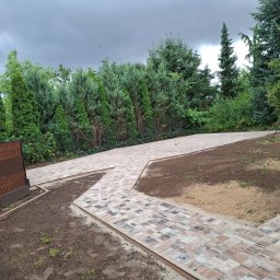 Piotr Wrzeszykowski - Kaskadowa ścieżka z kostki brukowej w ogrodzie, wykończona ozdobnymi obrzeżami. W tle zielone drzewa i krzewy, po prawej stronie osoba z narzędziem ogrodniczym.