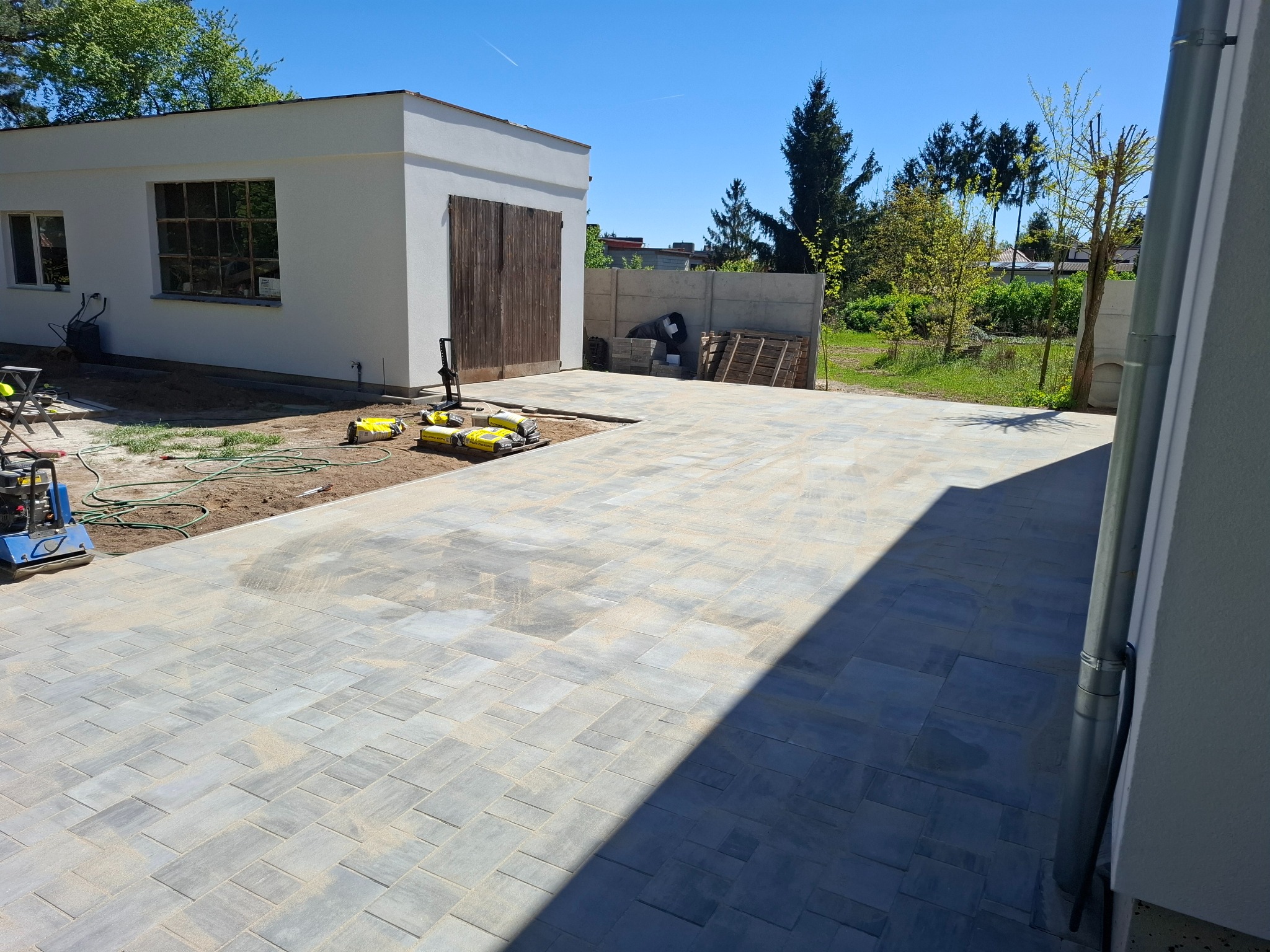 Jasnoszara kostka brukowa ułożona na podjeździe, obok garażu z drewnianymi wrotami i sprzętem budowlanym. Słoneczny dzień.