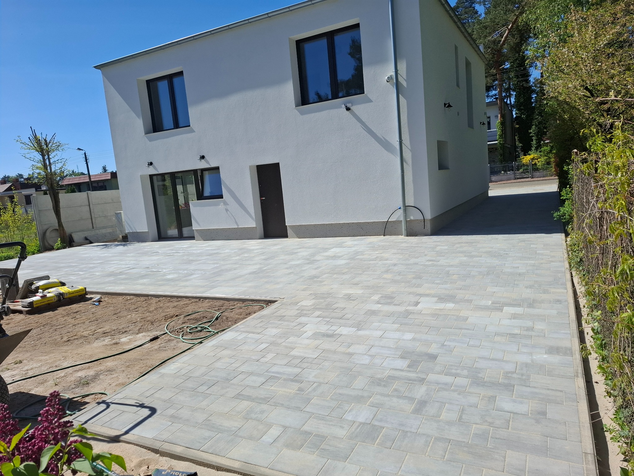 Jasnoszara kostka brukowa ułożona przed nowoczesnym, białym domem. Widoczne worki z materiałem i narzędzia brukarskie. Częściowo wykończony obszar z piaskiem.