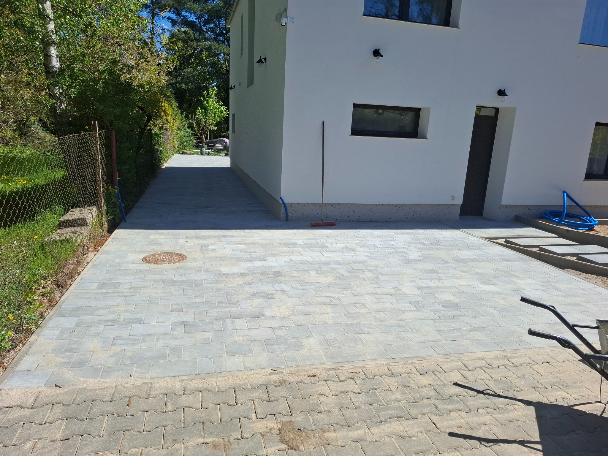 Nowa, szara kostka brukowa ułożona przed nowoczesnym, białym domem. Widoczny fragment starej nawierzchni i wózek transportowy.
