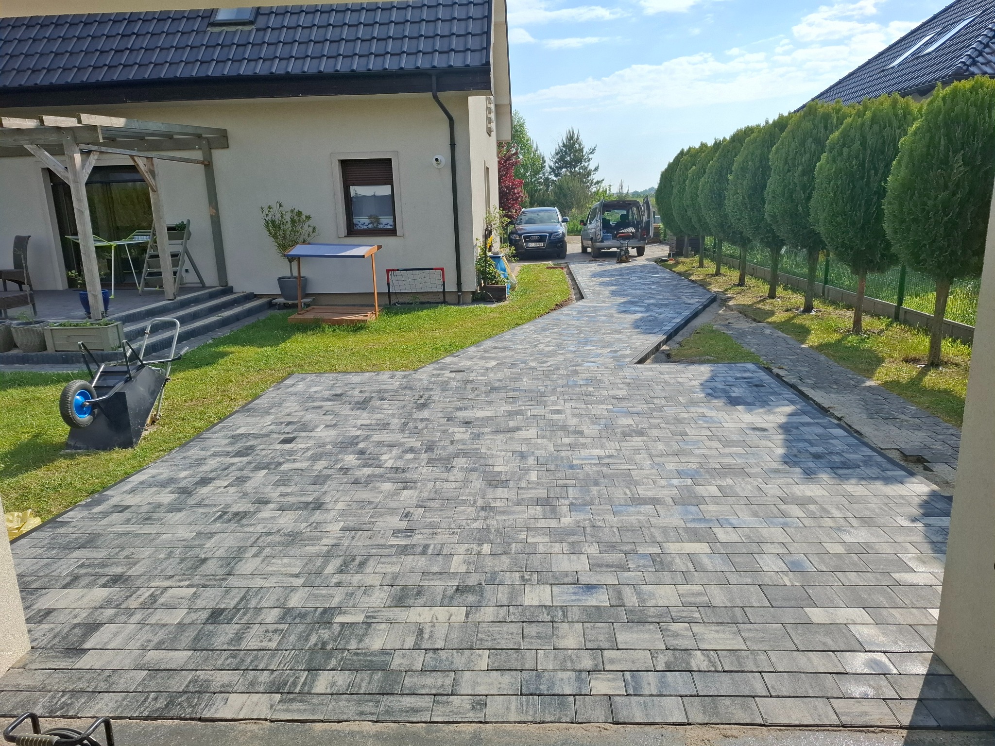 Szara kostka brukowa ułożona na podjeździe domu jednorodzinnego. W tle samochody i rząd tuj. Widok z perspektywy osoby stojącej na podwórku.