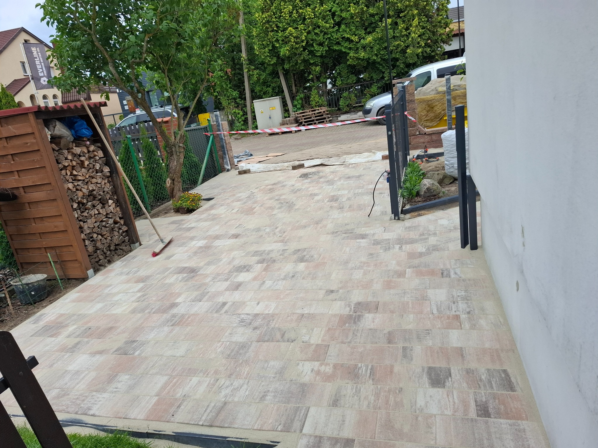Ułożona kostka brukowa w odcieniach beżu i szarości tworzy wzór na podjeździe. Widoczny fragment ogrodu, drewniana wiata na drewno i elementy ogrodzenia.