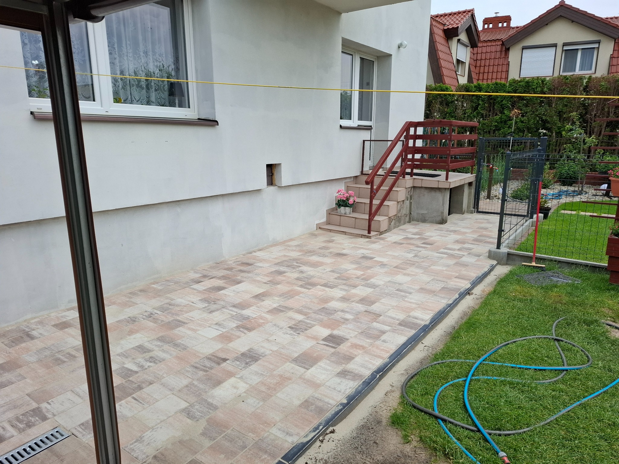 Wyłożony kostką taras przy domu z schodami i balustradą. Widoczny trawnik, płot i sąsiedni budynek. Kostka ułożona w oryginalny wzór.