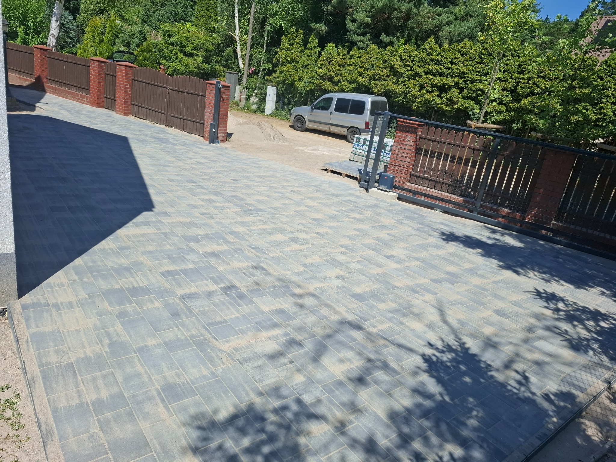 Ułożona kostka brukowa na podjeździe z ogrodzeniem z ceglanymi słupkami i drewnianymi panelami. W tle zaparkowany samochód i zieleń drzew.